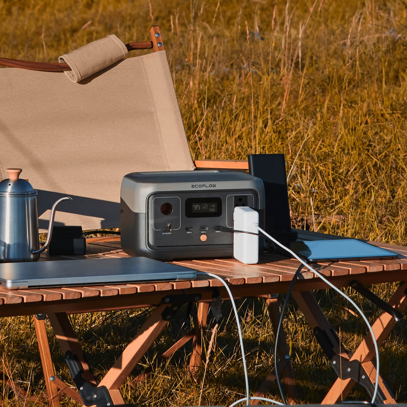 EcoFlow RIVER 2 on a wooden campsite table charging a laptop and a tablet, with a camping chair and kettle beside it