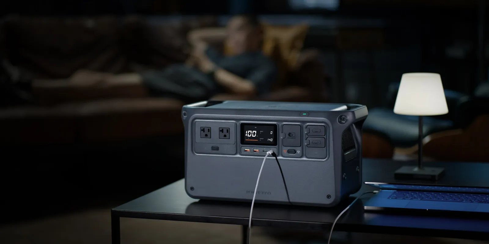 DJI Power 1000 portable power station on a table in a living room, front panel with two AC outlets, display and SDC ports visible, charging a laptop