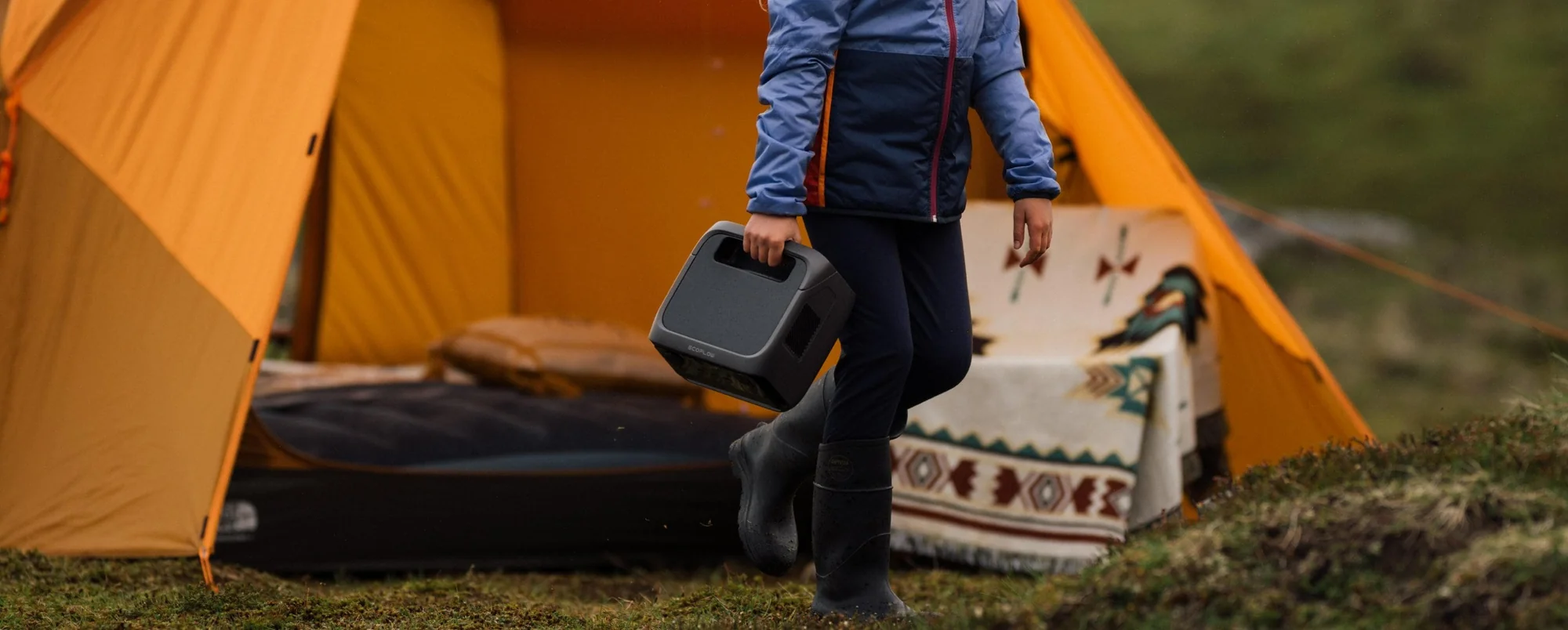 Person carrying the EcoFlow River 3 Plus at a campsite, showing its compact portable size